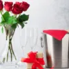 Elegant anniversary gift arrangement featuring a clear glass vase with red roses, two empty champagne flutes, a wrapped gift with a red ribbon, and a bottle of champagne in a stainless steel ice bucket with a red cloth, symbolizing romantic celebration and personalized gifts