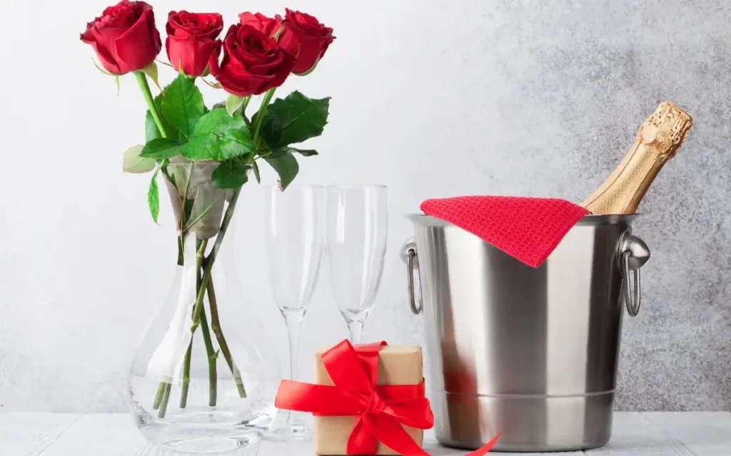 Elegant anniversary gift arrangement featuring a clear glass vase with red roses, two empty champagne flutes, a wrapped gift with a red ribbon, and a bottle of champagne in a stainless steel ice bucket with a red cloth, symbolizing romantic celebration and personalized gifts