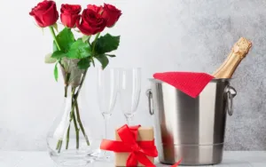 Elegant anniversary gift arrangement featuring a clear glass vase with red roses, two empty champagne flutes, a wrapped gift with a red ribbon, and a bottle of champagne in a stainless steel ice bucket with a red cloth, symbolizing romantic celebration and personalized gifts